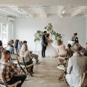A small gathering witnesses a beautiful wedding ceremony indoors with wooden chairs and floral decor.